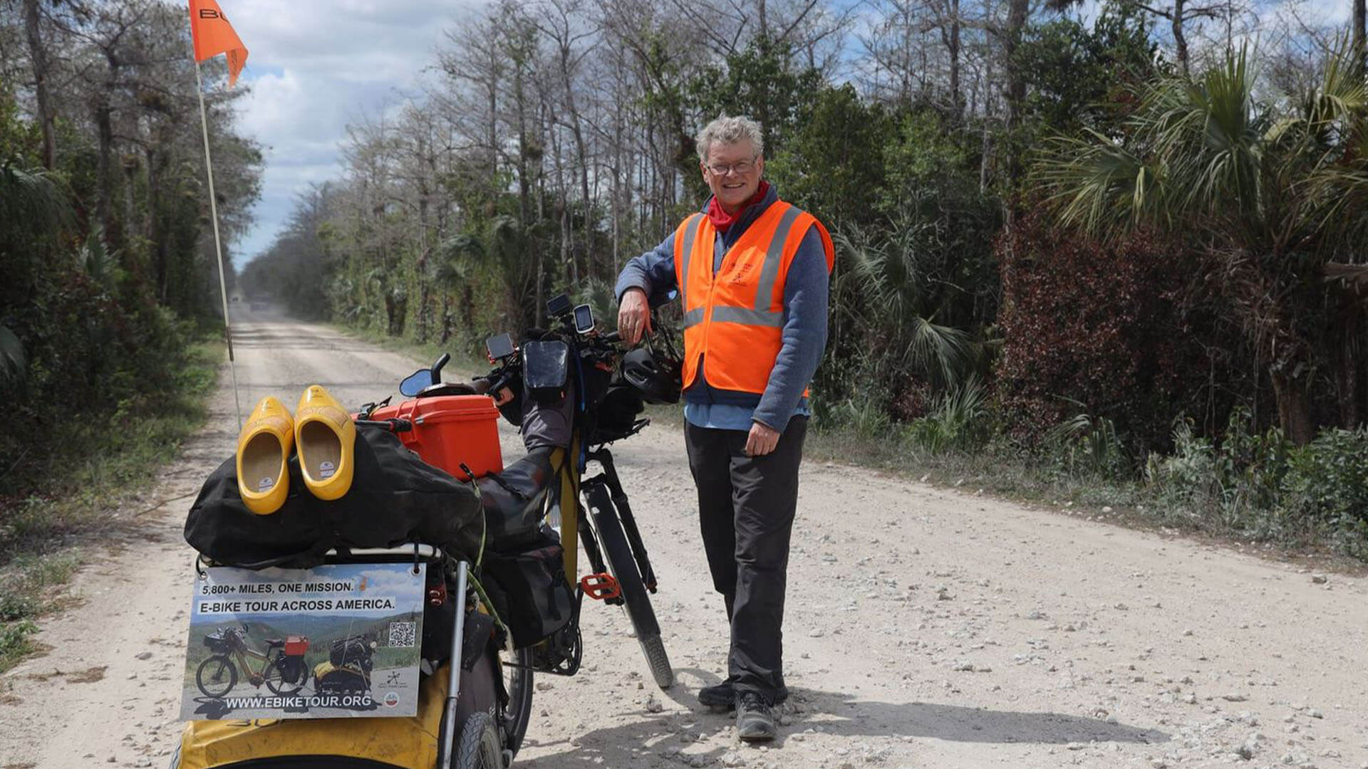Person standing beside a loaded e‑bike on a rural dirt road, with orange safety vest and gear for a cross‑country bike tour visible on the bicycle.