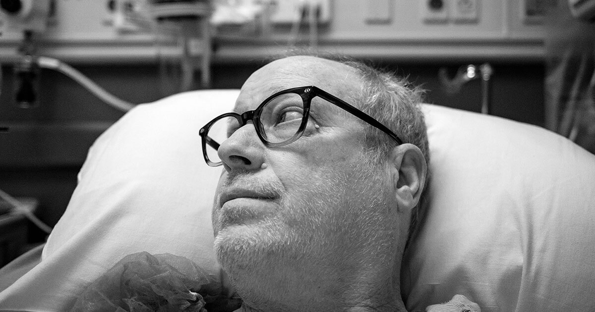 Person wearing glasses lying on a hospital bed, looking upward, with medical equipment blurred in the background.