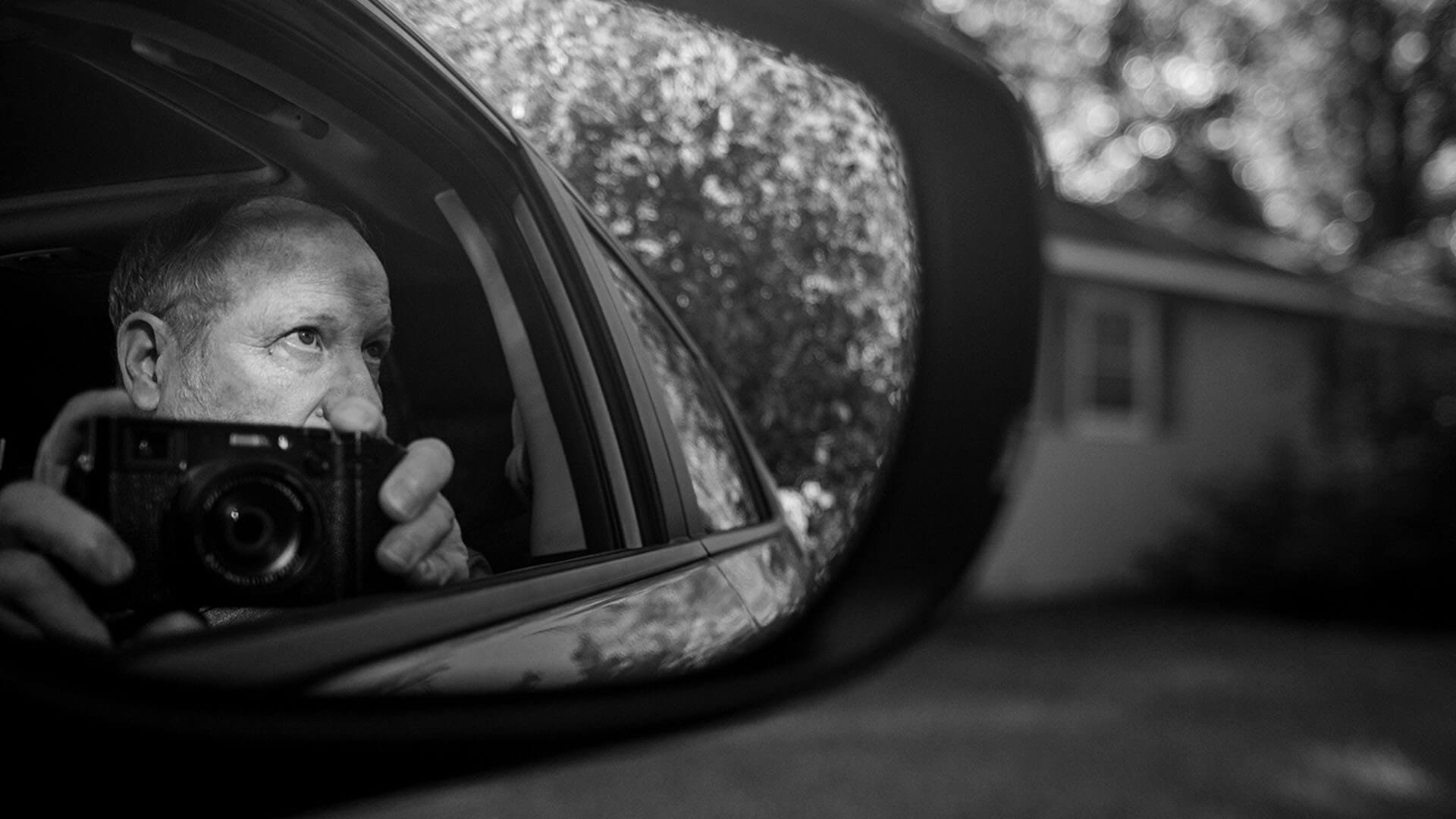 Man holding a camera reflected in a car side mirror, with a blurred residential street in the background.