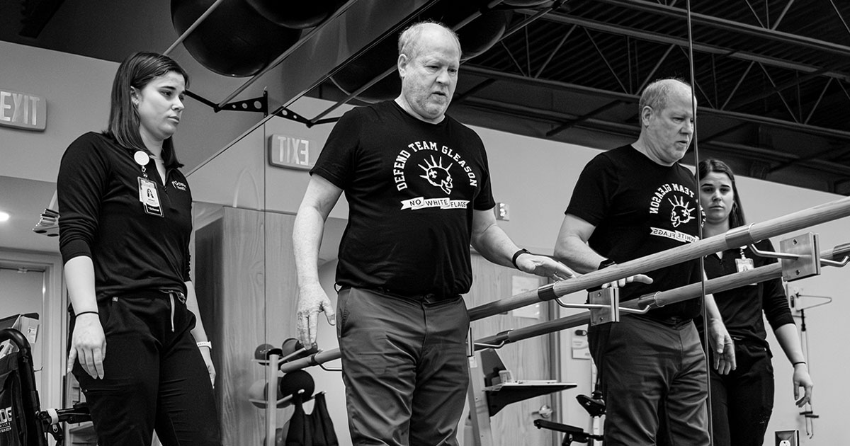 Individual performing gait training in a rehabilitation facility, walking between parallel bars with assistance from two clinicians.