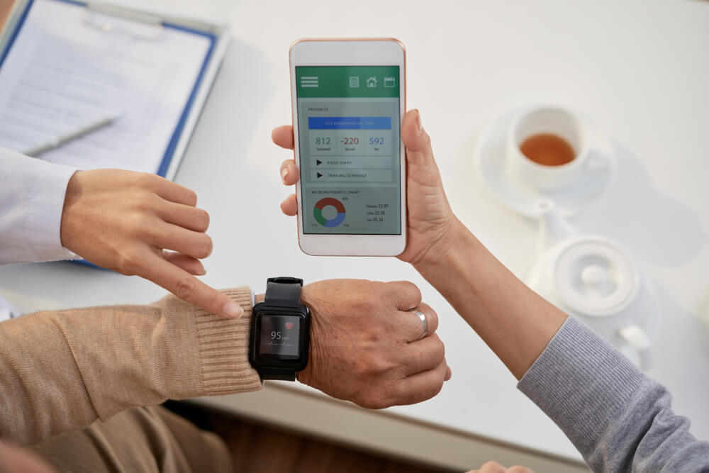 Young physician wearing white coat explaining senior patients how to use mobile application in order to control heart rate while having consultation at office