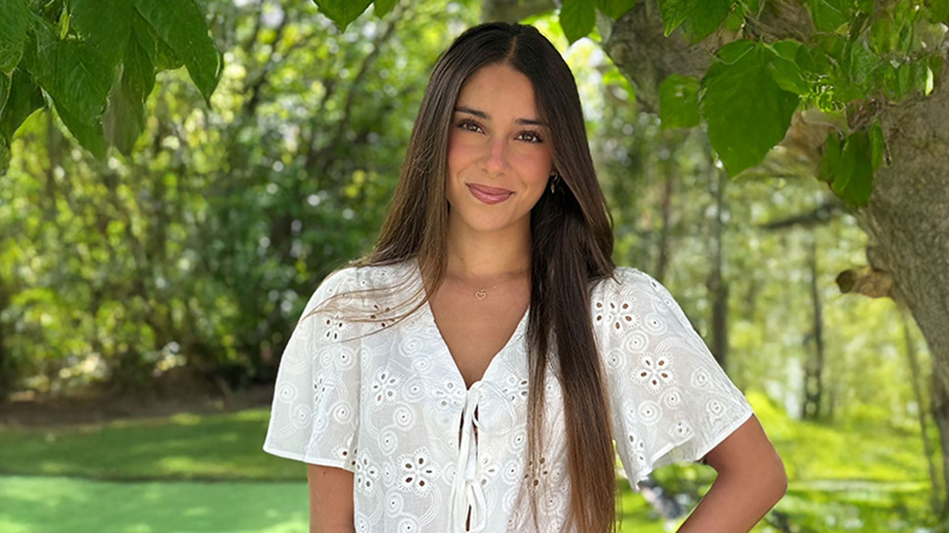 Portrait of a person standing outdoors beneath a tree, wearing a white blouse, with green foliage in the background.