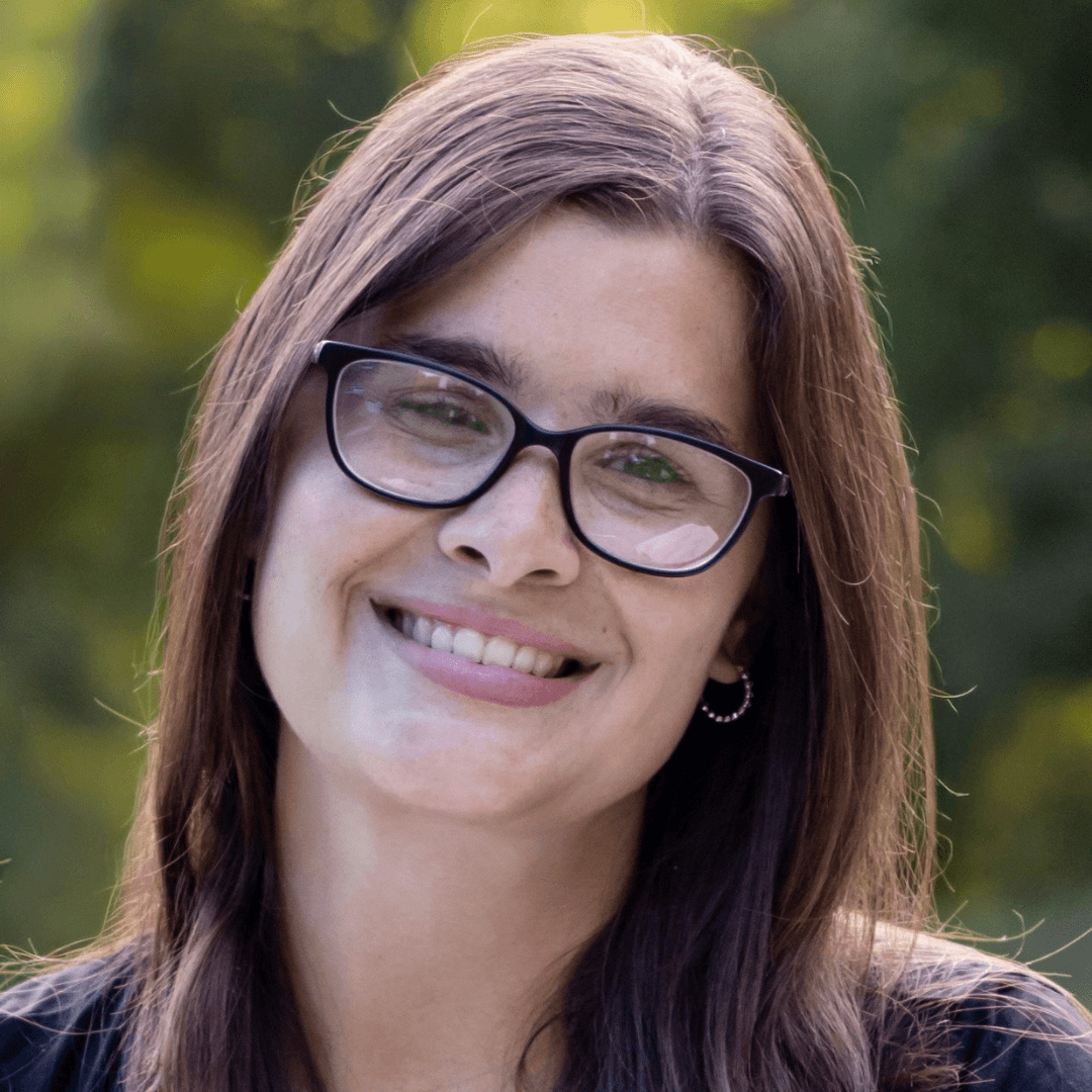 Smiling woman with long brown hair and glasses outdoors with a blurred green background.