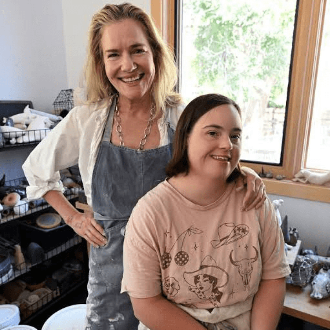 Two people posed together in a pottery studio, one standing behind with an arm on the other’s shoulder, shelves of ceramics and tools in the background.
