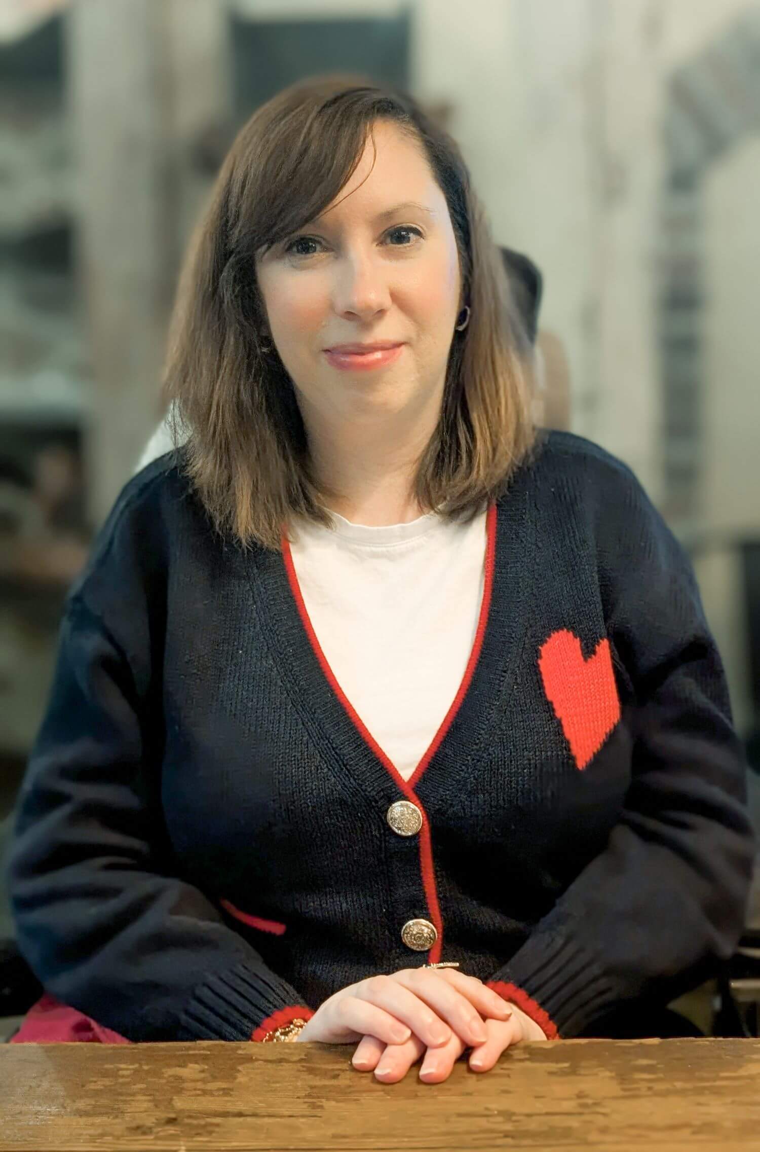 Portrait of Victoria Nedza seated at a table, facing the camera, wearing a dark cardigan with red trim over a light shirt, with a softly blurred indoor background.