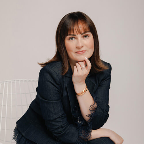 Professional headshot of Molly Burke wearing a dark blazer, seated and resting her chin on her hand against a neutral background.