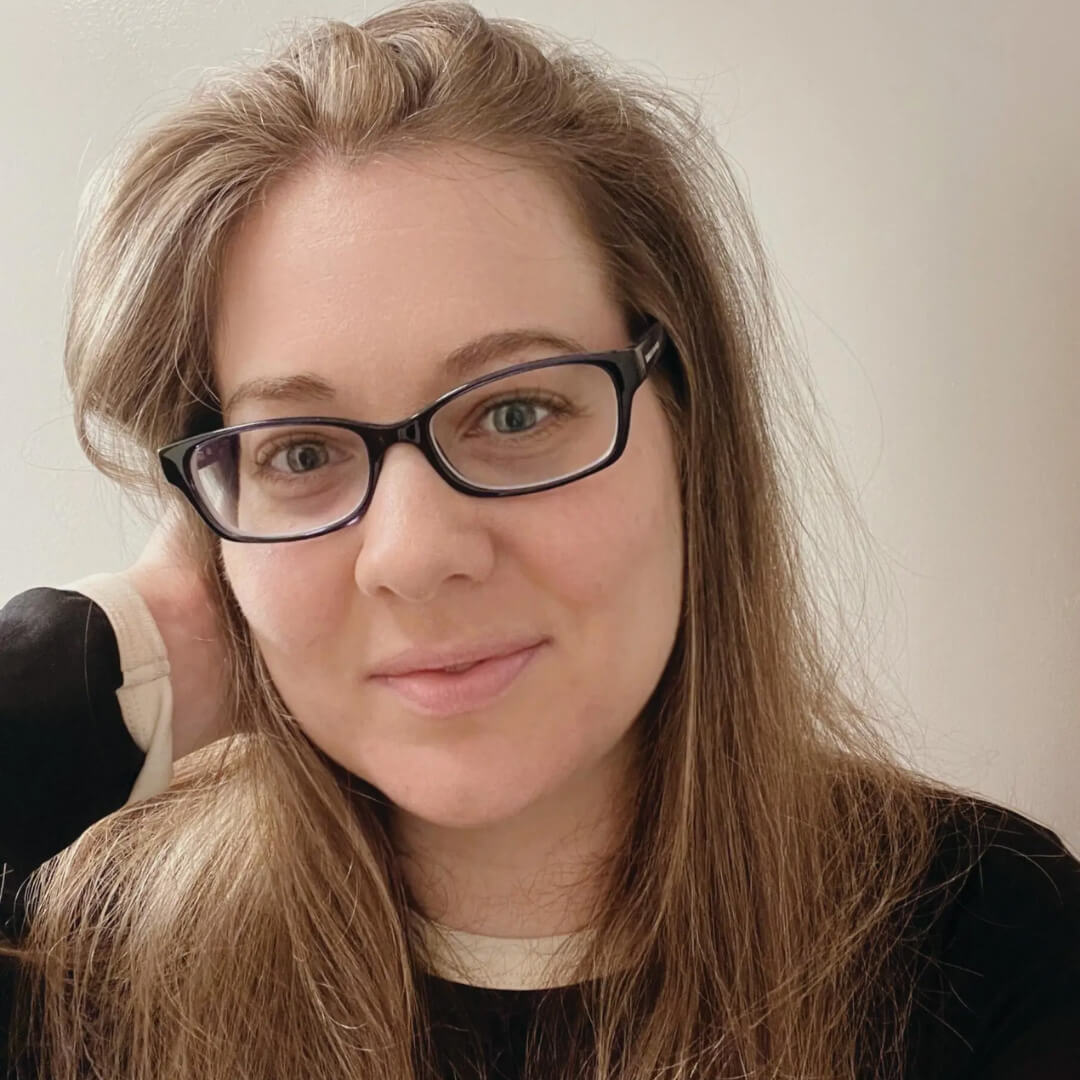 Portrait of a person with long light brown hair wearing black rectangular glasses, resting their head on one hand, against a neutral indoor background.