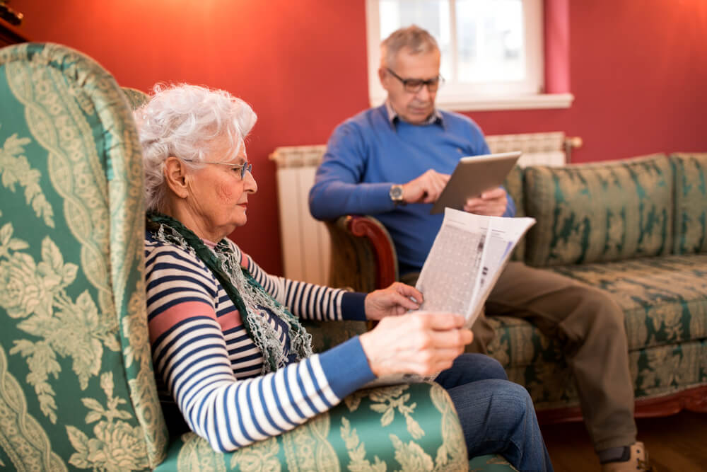Senior lovely couple relax at home while reading newspaper and using tablet