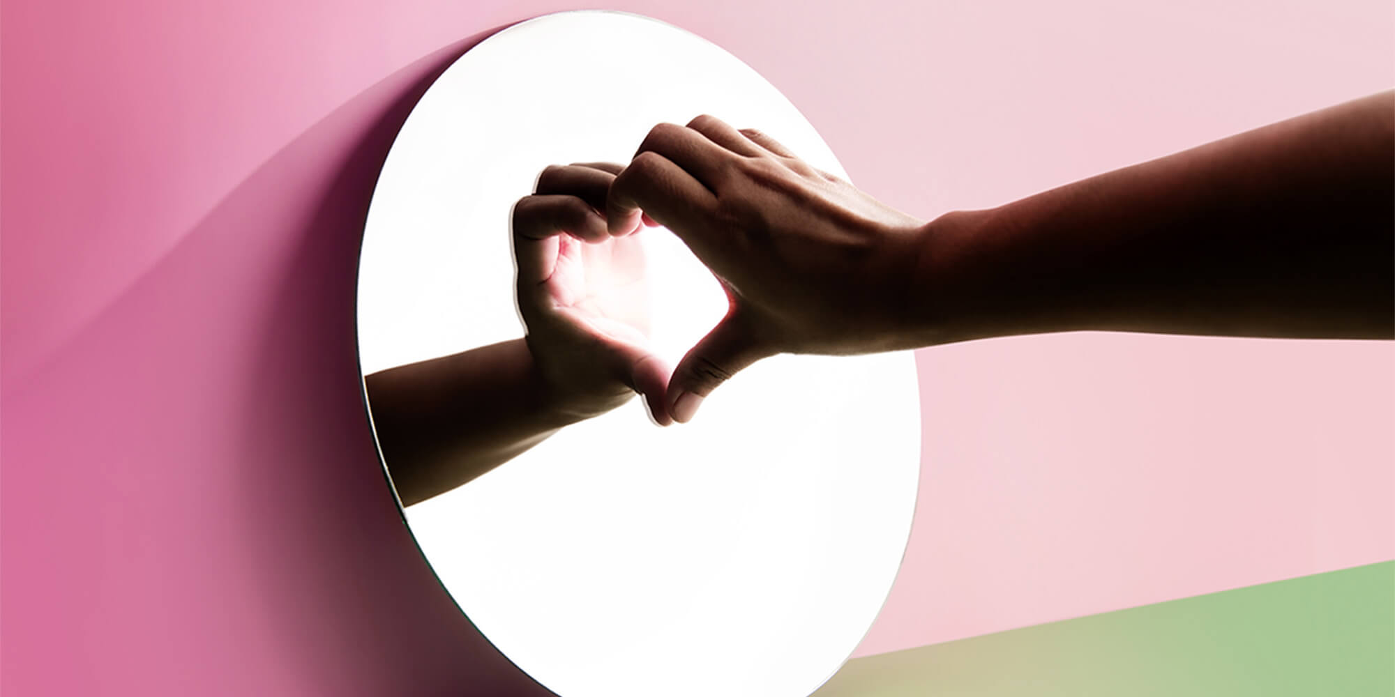 Hands forming a heart shape through a circular opening against a pink background, symbolizing care and self-love.