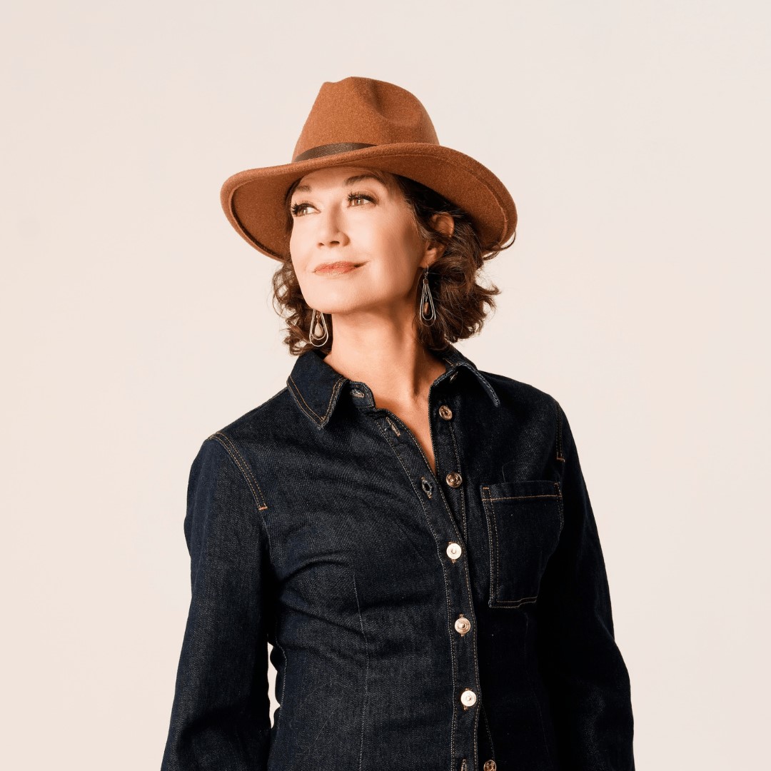 Portrait of a person wearing a wide-brim tan hat, dangling earrings, and a dark button-up denim shirt, looking slightly upward against a neutral background.