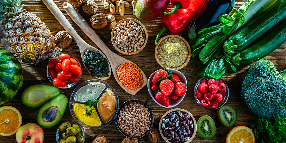 A colorful assortment of Mediterranean diet foods on a wooden table, including fruits, vegetables, legumes, nuts, seeds, olive oil, and whole grains arranged in bowls and spoons.