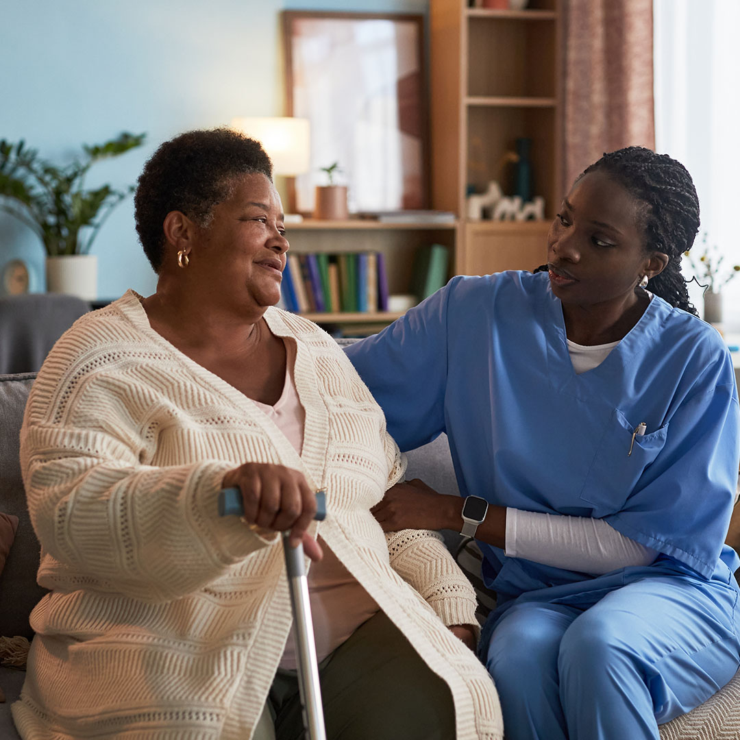 Senior Black woman sitting on sofa holding cane, talking with young adult Black female nurse in medical scrubs, providing support and companionship in home setting.