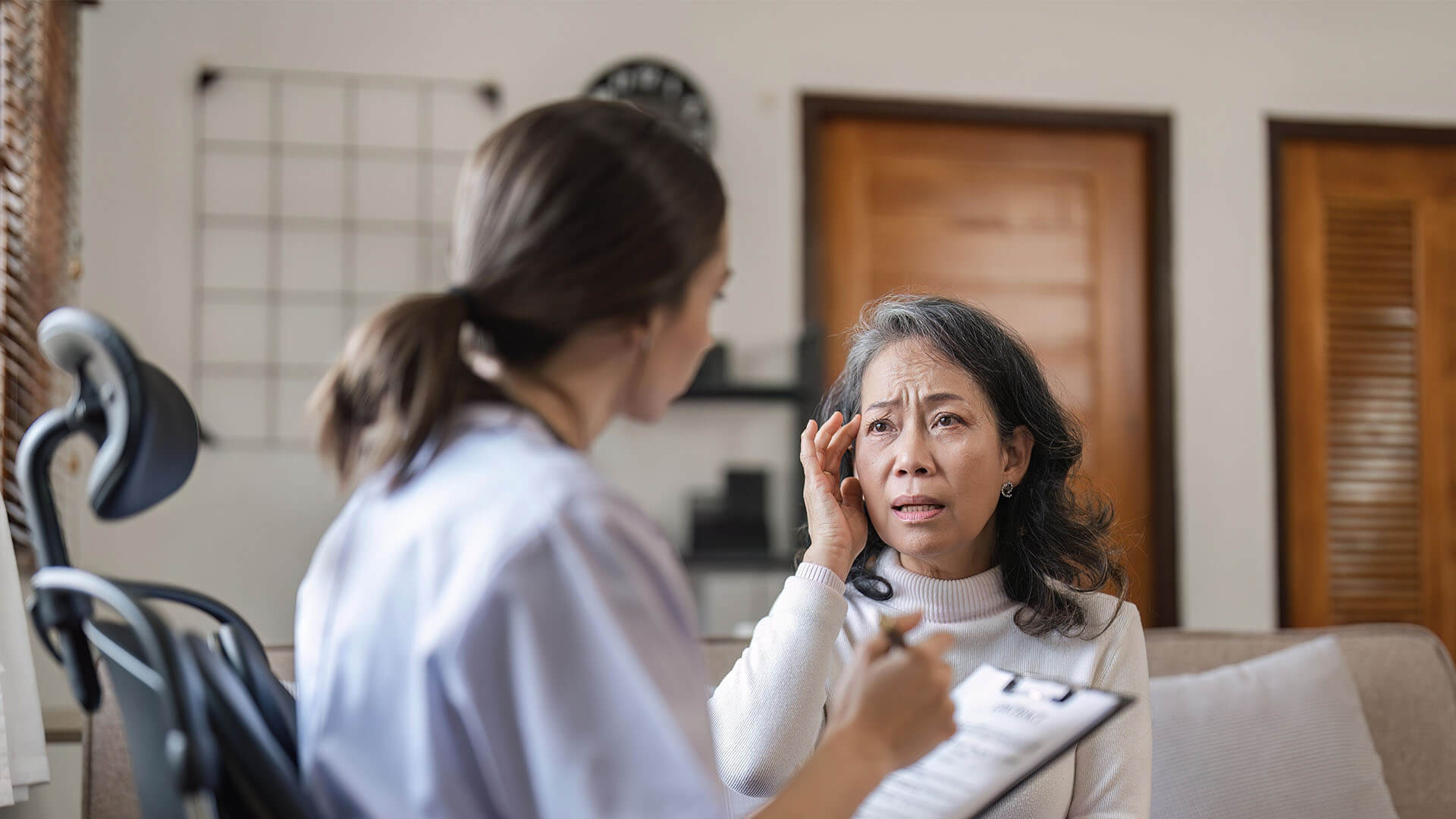 Female doctor is examining elderly patient while taking notes of illness. Patient is having headache.