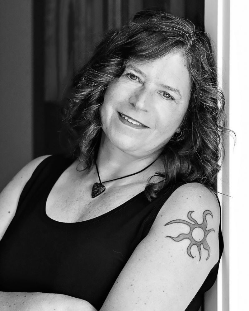 Black-and-white portrait of a woman smiling, leaning against a wall, with a sun tattoo visible on her upper arm.