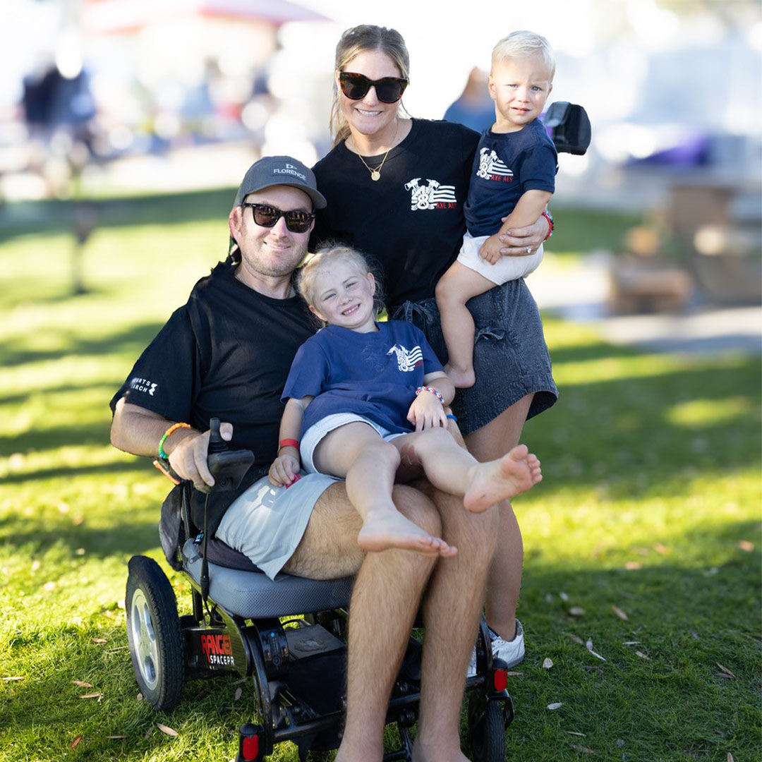 Family of four wearing matching ALS awareness shirts poses outdoors; father uses a powered wheelchair while holding his daughter, with mother holding a toddler beside him.