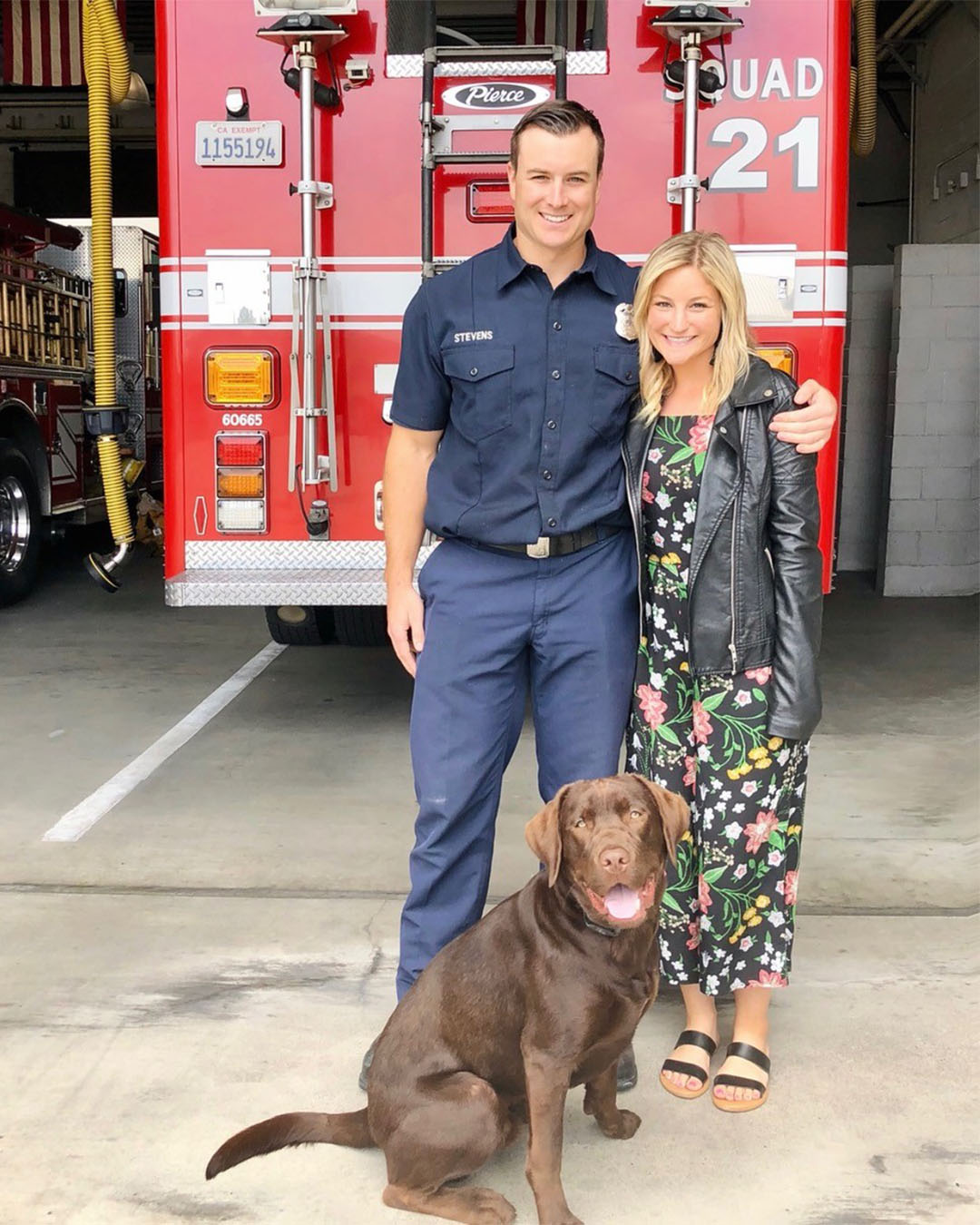 Firefighter Eric Stevens and his wife pose with their dog in front of a fire engine at a fire station.