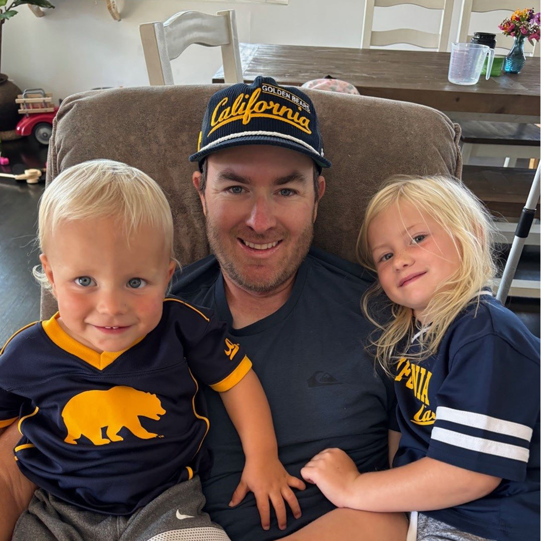 Eric Stevens sits with his two young children at home, all wearing University of California Golden Bears apparel and smiling for the camera.