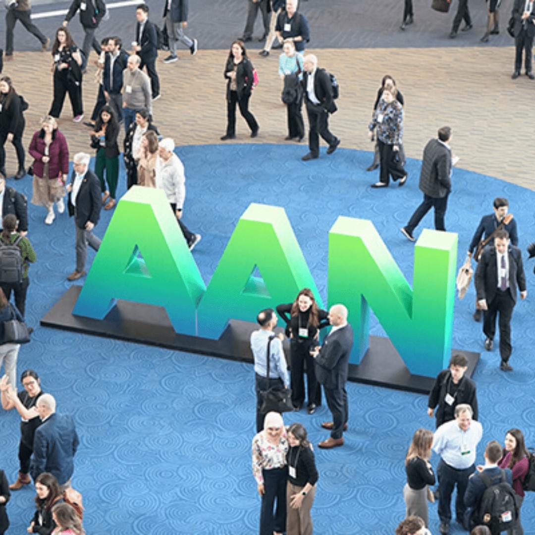 People walking and talking around a large green “AAN” sign on a blue carpeted floor at a conference or event space.