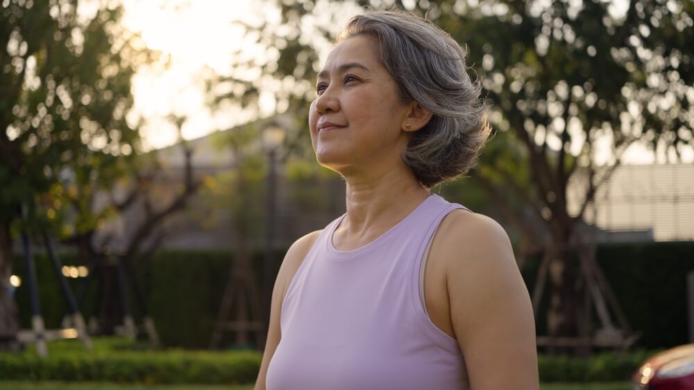 Older Asian woman practicing yoga with arms open in a sunny park, promoting relaxation, mindfulness, and healthy aging.
