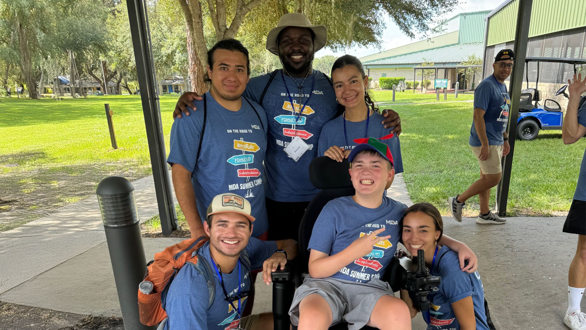 Group of MDA Summer Camp counselors and a smiling camper in a wheelchair outdoors under trees