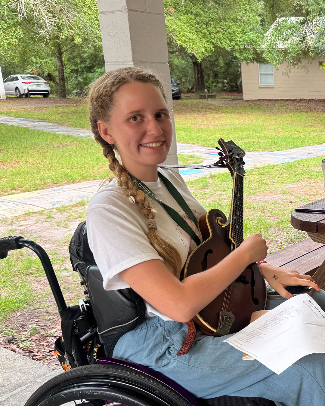 Woman in a wheelchair smiling while holding a mandolin and sheet music at an outdoor camp pavilion