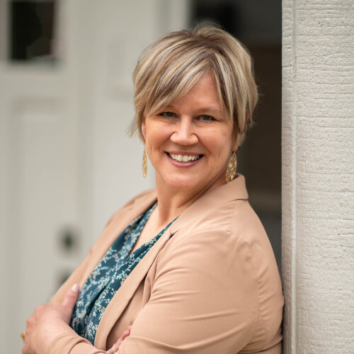 Portrait of Lori Jones standing with arms crossed, wearing a light blazer over a patterned top, leaning against a neutral wall.