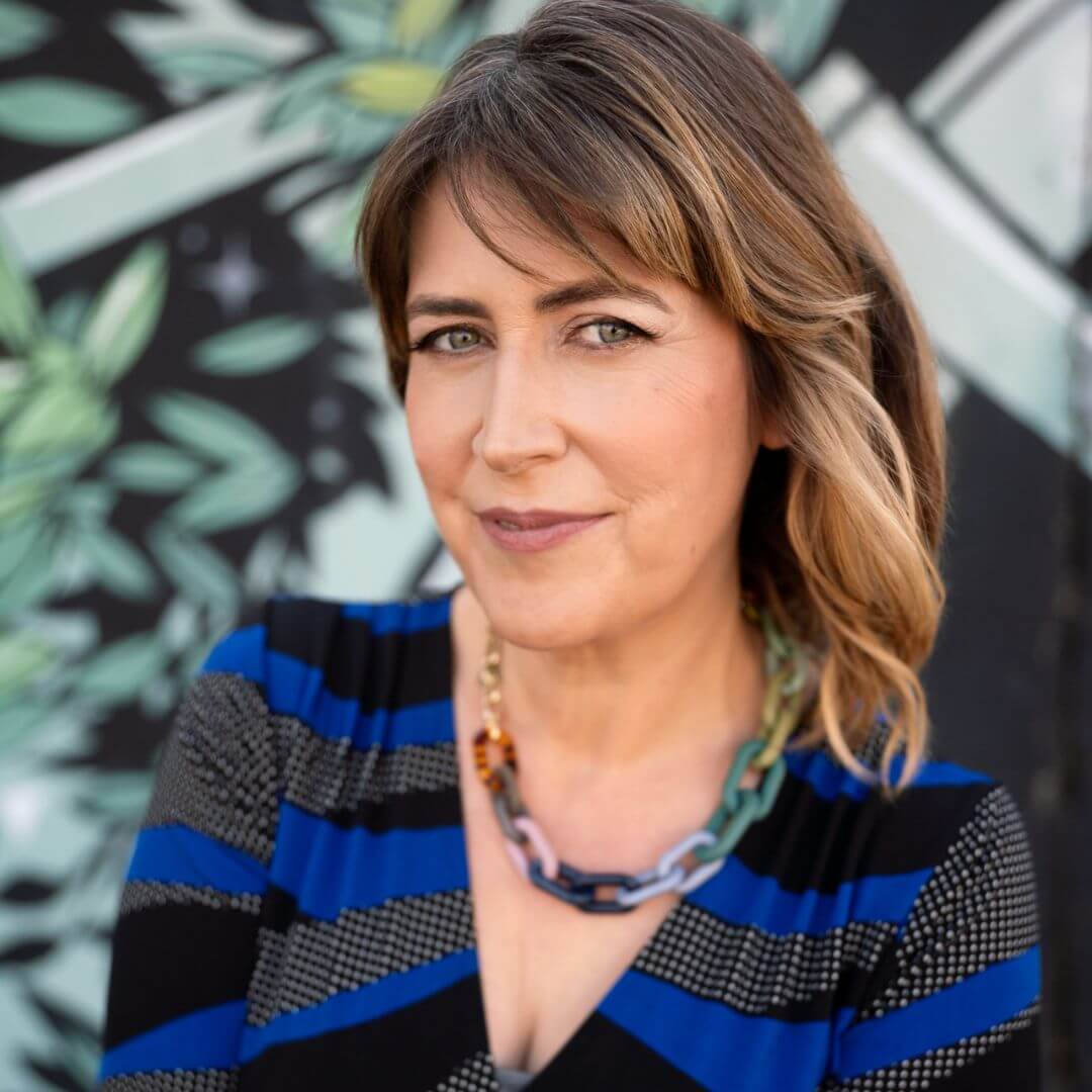 Portrait of Darby Saxbe smiling, wearing a blue and black patterned top and necklace, photographed against a leafy mural background.