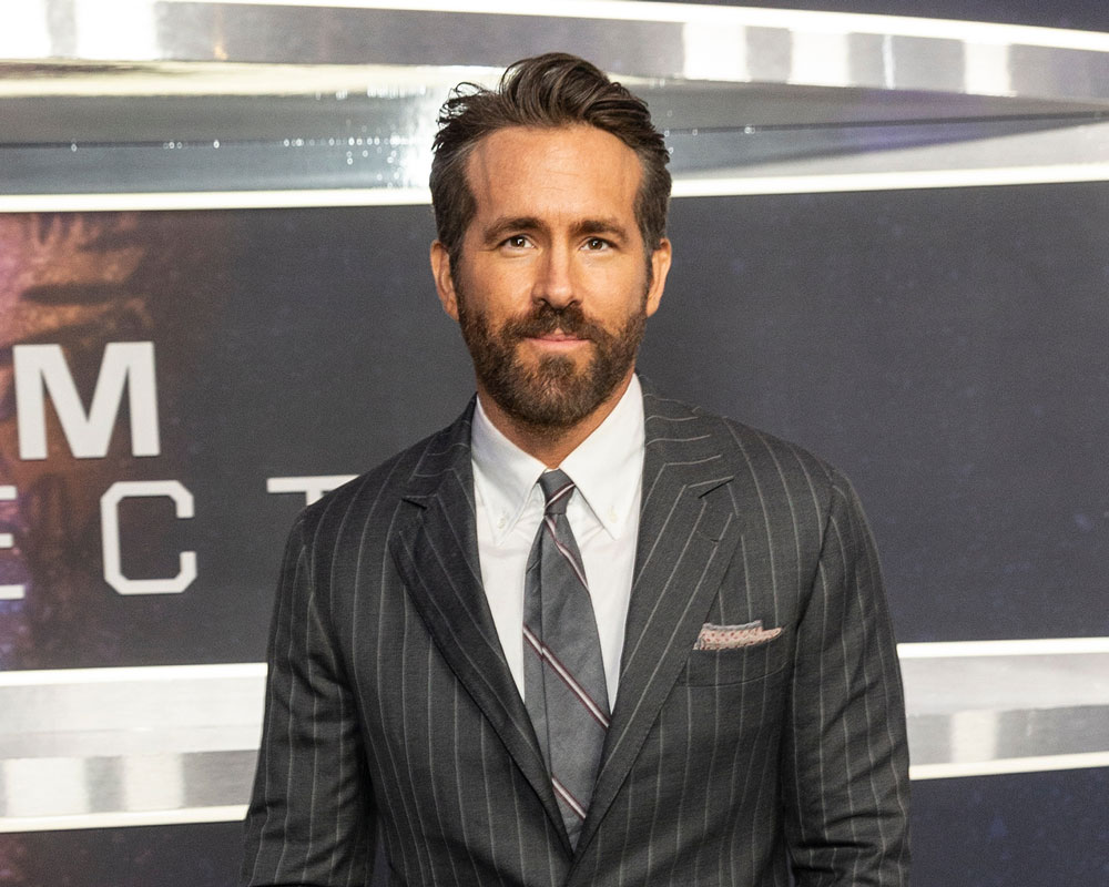 Ryan Reynolds with dark hair and a beard wearing a dark pinstripe suit, white shirt, and striped tie at a formal event.