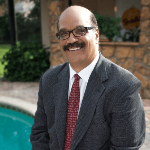 Mark McEwen seated in a dark gray suit and red tie beside a pool, smiling in a relaxed upscale setting.