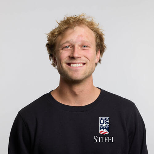 Colby Stevenson smiling with light blond, slightly curly hair wearing a black shirt with a U.S. Ski Team logo on the chest, standing against a plain light gray background.
