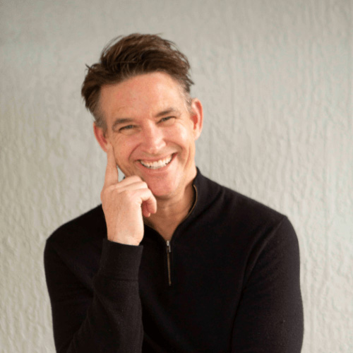 Rob Floyd smiles at the camera, wearing a black long-sleeve top, with his hand resting against his face against a light-colored background.