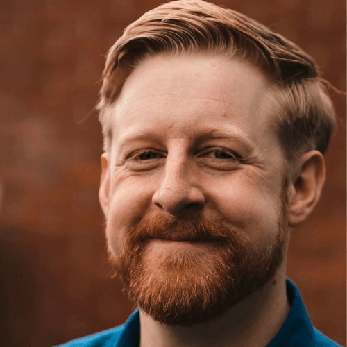 Headshot of Michael Patrick, an Irish actor and writer, smiling, with short hair and a beard, against a brick background.